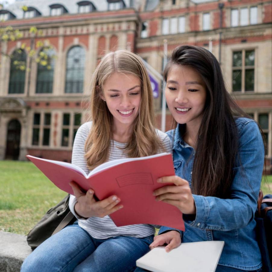 Two female students sitting on steps outside a building, engaged in conversation and studying.