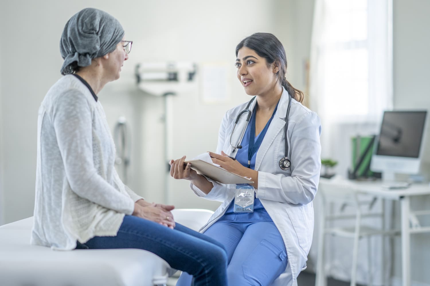 A female Cancer patient sits up on an exam table during a check-up. She is dressed comfortably and has a headscarf on to keep warm as she talks with her Oncologist. The doctor is wearing a white lab coat and taking notes on a clipboard.