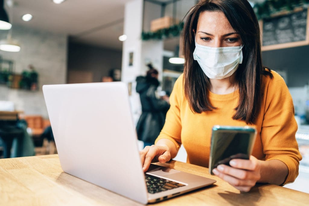 Young woman using laptop and mobile phone at cafe wearing face mask