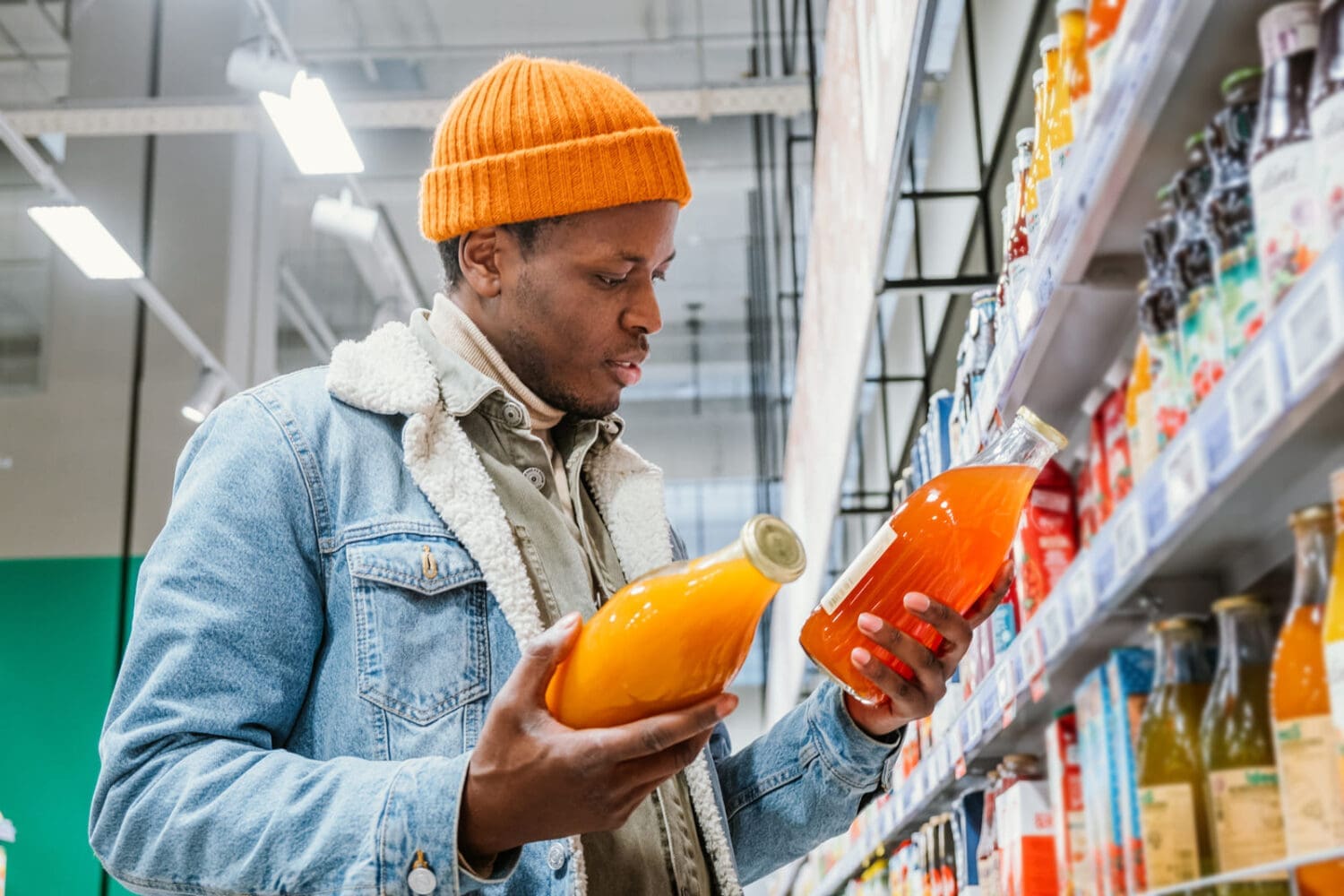 African man in a stylish orange hat and denim jacket chooses natural juice in glass bottles in a grocery supermarket standing at the shelves with products - William Russell blog - How Can Expats And Digital Nomads Survive Cost Of Living Crisis