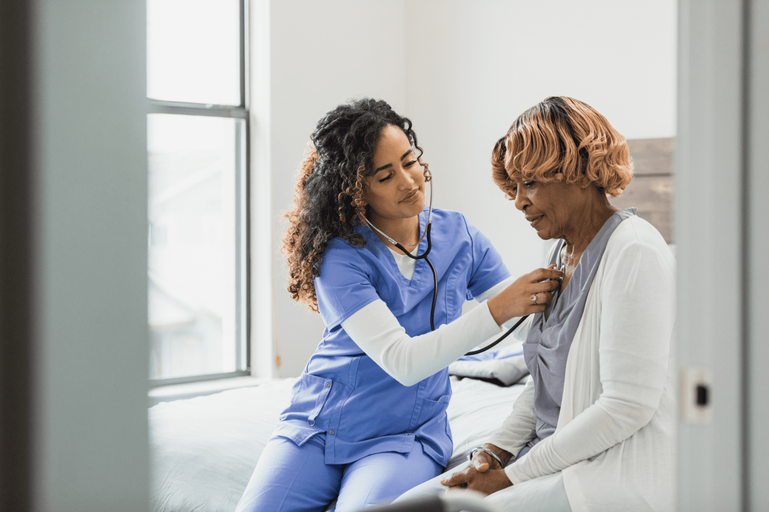 Female nurse sitting beside a senior female patient using a stethoscope to check her heart