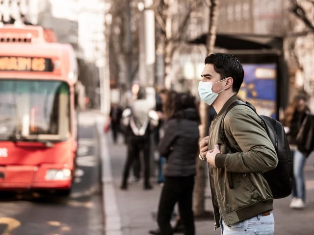 A young man wearing a face mask stands at a bus stop
