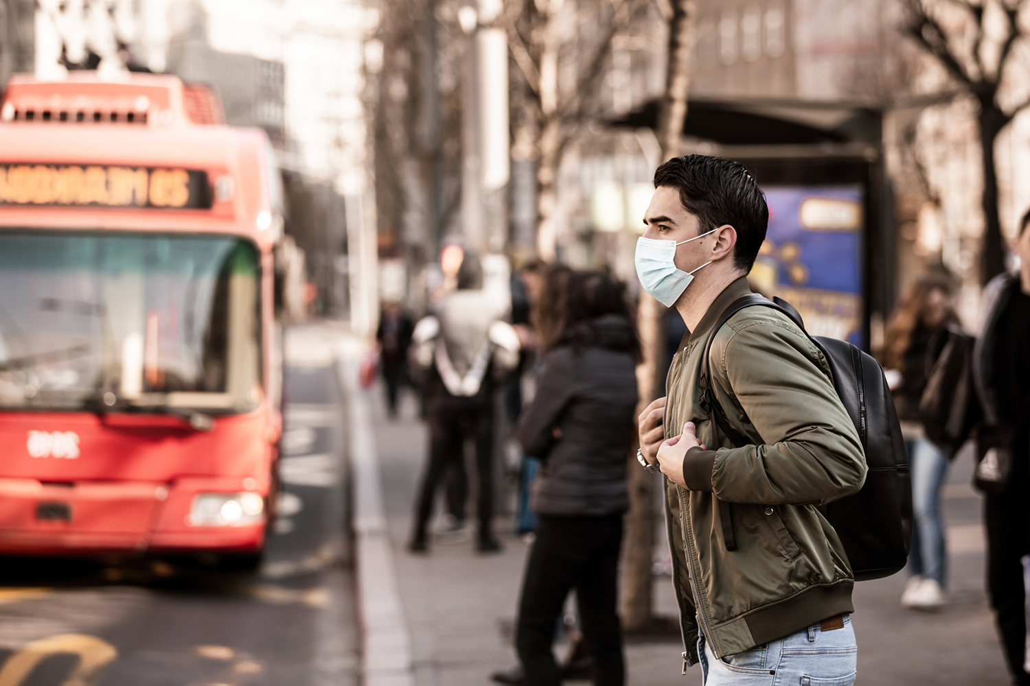 A young man wearing a face mask stands at a bus stop