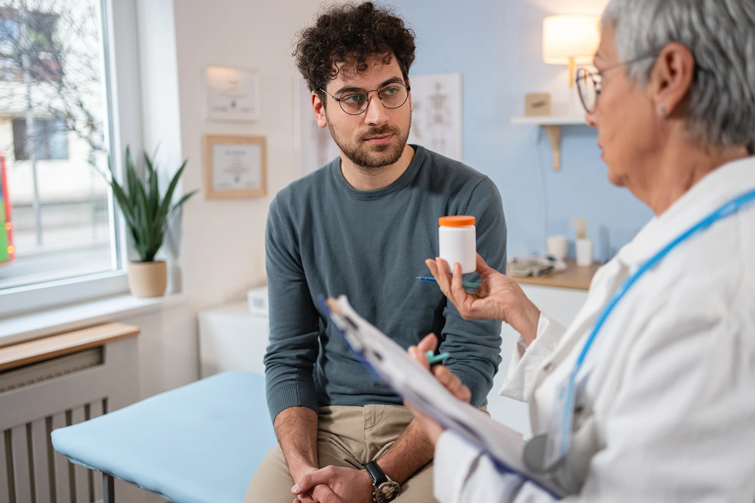 Senior female doctor standing talking to a male patient who is sitting on a bed at the doctors
