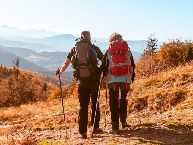 A couple out hiking together with walking poles and large rucksacks on their backs, facing out onto mountains in the distance