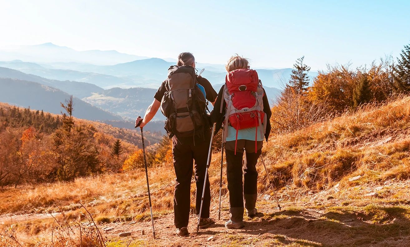 A couple out hiking together with walking poles and large rucksacks on their backs, facing out onto mountains in the distance